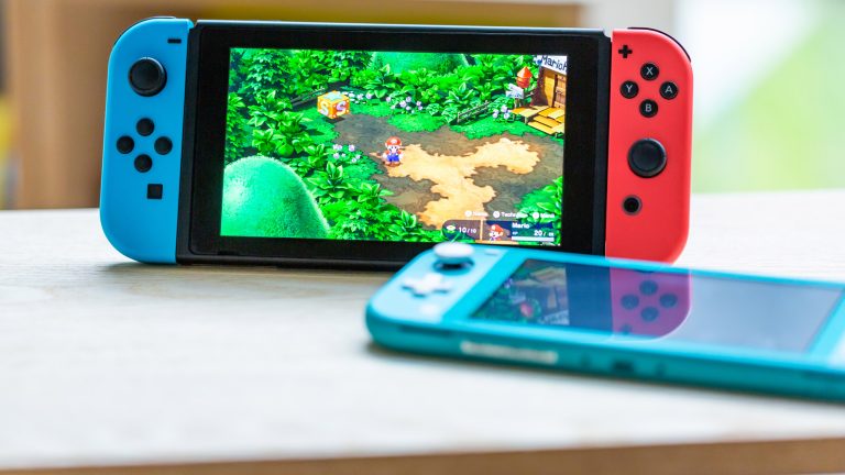 Eine Nintendo Switch steht auf einem Tisch, daneben liegt eine Nintendo Switch Lite.