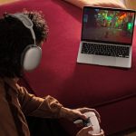 Eine Person hockt vor einem MacBook Air und h&auml;lt dabei einen Controller in der Hand. Auf dem MacBook l&auml;uft ein Spiel.