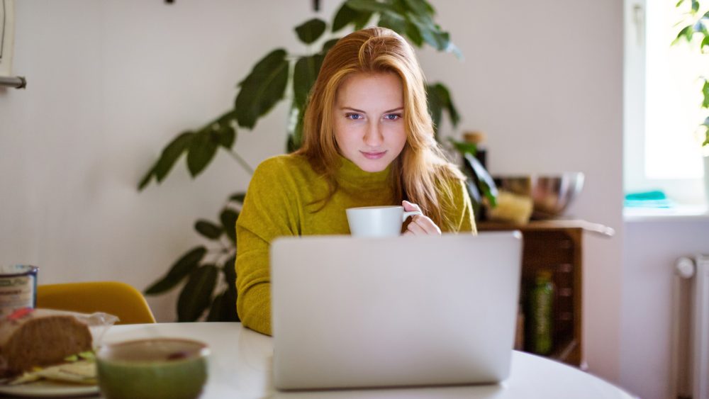 Eine Person mit einer Kaffeetasse in der Hand sitzt vor einem Laptop.