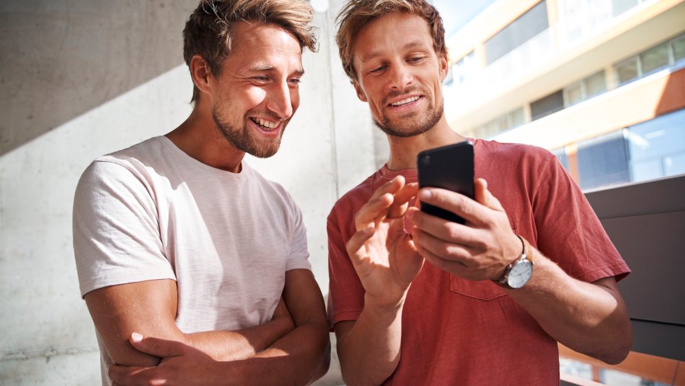 Zwei Personen stehen nebeneinander und lachen zusammen in ein Smartphone.