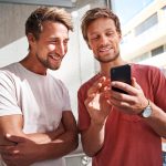 Zwei Personen stehen nebeneinander und lachen zusammen in ein Smartphone.