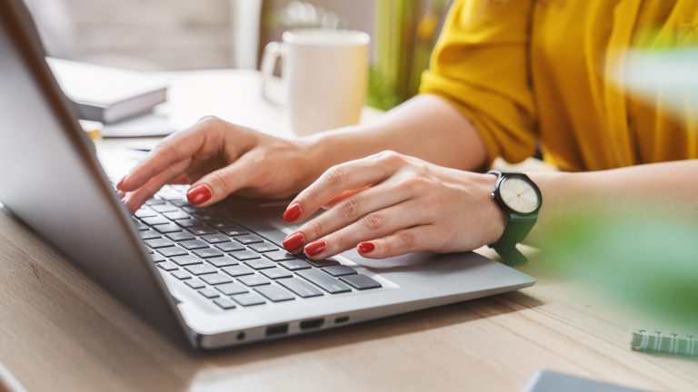 Eine Person tippt mit beiden Händen auf ihrer Laptoptastatur. Sie trägt eine gelbe Bluse, eine schwarze Armbanduhr und roten Nagellack.