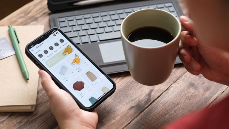 Eine Person mit Kaffee in der einen Hand, hält ein Smartphone mit geöffnetem Online-Shop in der anderen.