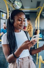 Eine Person mit vielen langen schwarzen Zöpfen steht im Bus und hört mit Over Ear Kopfhörern Musik von ihrem Handy.