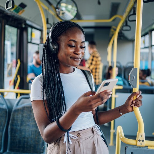 Eine Person mit vielen langen schwarzen Zöpfen steht im Bus und hört mit Over Ear Kopfhörern Musik von ihrem Handy.