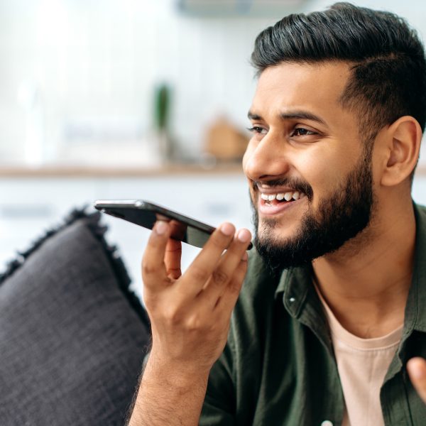 Ein Mensch sitzt in einem dunkelgr&uuml;nen Hemd auf einer grauen Couch und telefoniert mit seinem iPhone in der Hand vor dem Mund.
