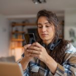 Eine Person sitzt an einem Tisch mit einem Laptop vor sich. Sie h&auml;lt ein Smartphone in der Hand.