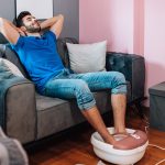 Ein Mensch sitzt mit hochgekrempelter Jeans auf einer grauen Couch. Die F&uuml;&szlig;e sind in einem wei&szlig;-ros&eacute;-farbenen und mit Wasser gef&uuml;llten Fu&szlig;massageger&auml;t.