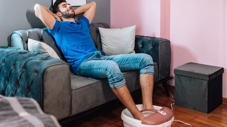Ein Mensch sitzt mit hochgekrempelter Jeans auf einer grauen Couch. Die F&uuml;&szlig;e sind in einem wei&szlig;-ros&eacute;-farbenen und mit Wasser gef&uuml;llten Fu&szlig;massageger&auml;t.