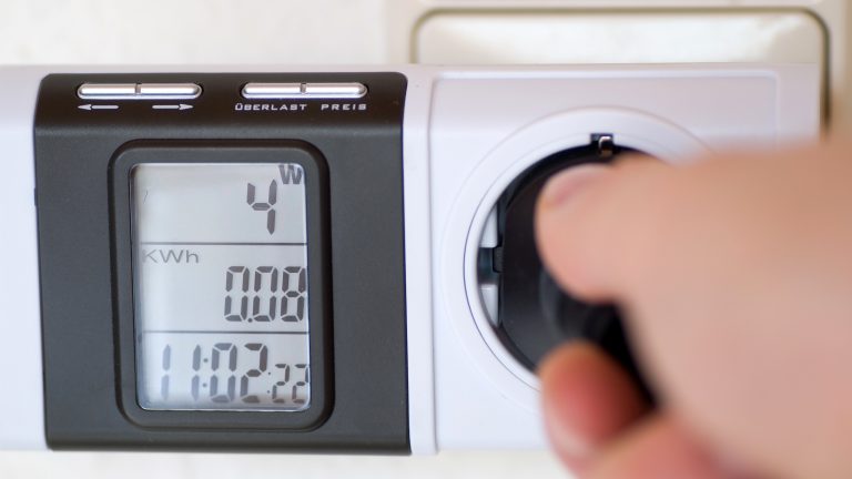 Ein Strommessgerät, das vor eine Steckdose gesteckt wird und den Stromverbrauch von vier Watt und 0,08 KWh anzeigt. Rechts eine Hand, die einen Stecker in diese Steckdose steckt.