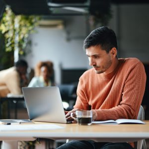 Ein Mensch in orangenem Pullover sitzt in einem Büro vor einem Laptop. Im Hintergrund sieht man andere Mitarbeitende und Pflanzen.