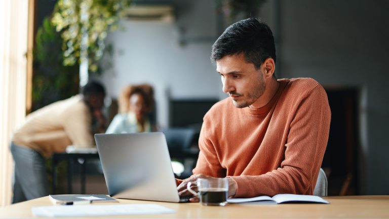 Ein Mensch in orangenem Pullover sitzt in einem Büro vor einem Laptop. Im Hintergrund sieht man andere Mitarbeitende und Pflanzen.