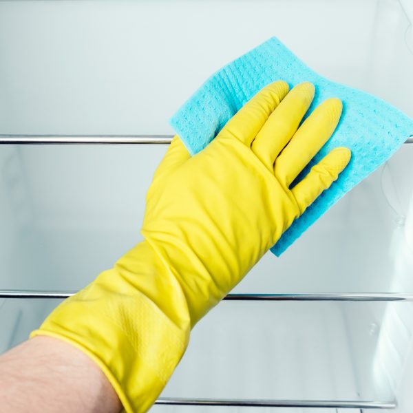 Eine Hand in einem gelben Gummihandschuh wischt die Ablageflächen in einem Kühlschrank mit einem blauen Schwammtuch. 
