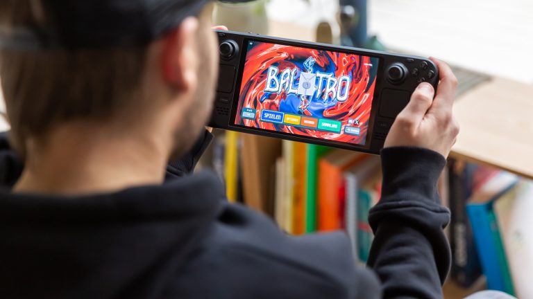 Eine Person schaut auf ein Steam Deck, das sie in der Hand hält. Darauf ist der Startbildschirm von Balatro zu sehen.