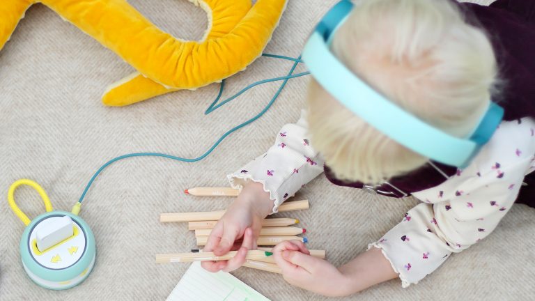 Ein Kind mit Kopfhörern auf den Ohren liegt auf dem Fußboden und ist mit Buntstiften beschäftigt, die vor ihm liegen. Daneben liegt der Player des Galakto, an dem der Kopfhörer angeschlossen ist.