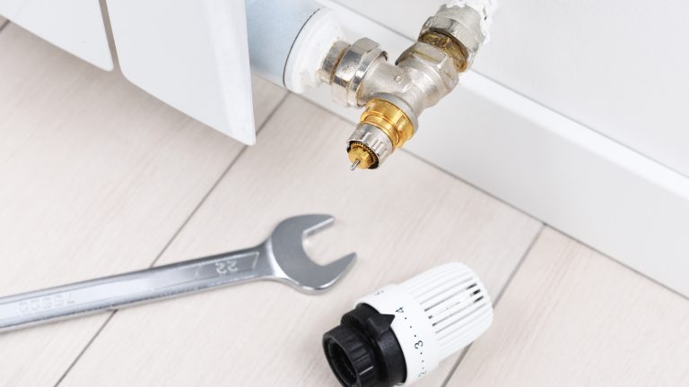 Neben einer Heizung mit abgeschraubtem Thermostat liegen ein Schraubenschlüssel und das abgebaute Teil auf dem Fußboden.