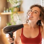 Eine Person mit halblangen, braunen Locken und rotem Tanktop f&ouml;hnt sich im Bad die Haare.