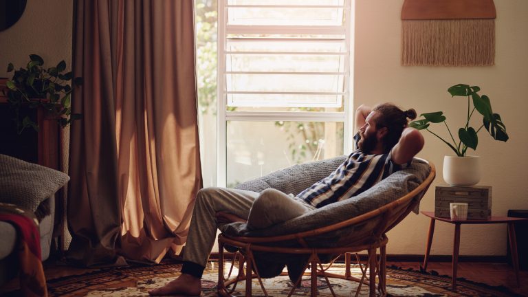 Eine Person sitzt mittig in einem Wohnzimmer auf einem runden Sessel, der auf einem Teppich steht, und genießt die Musik. Vor dem Fenster sind dunkelrote, schwere Vorhänge.