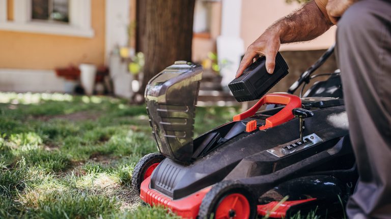 Eine Person knieht neben einem schwarz-roten Akkurasenm&auml;her und ist im Begriff den schwarzen Akku mit der rechten Hand in den M&auml;her zu legen.