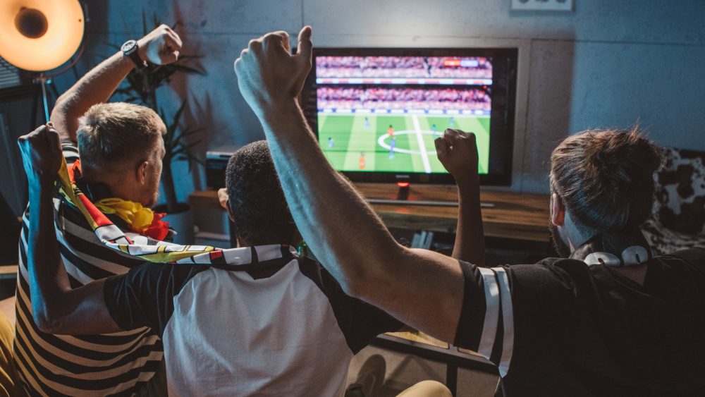 Drei Personen sitzen vor einem Fernseher und schauen ein Fußballspiel.