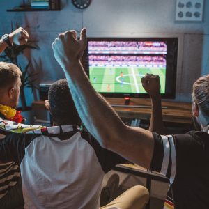 Drei Personen sitzen vor einem Fernseher und schauen ein Fu&szlig;ballspiel.