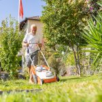 Ein Mensch mit wei&szlig;em T-Shirt m&auml;ht mit einem wei&szlig;-orangenen Akkurasenm&auml;her seinen Garten. Die Sonne scheint und der Rasen und die Pflanzen sind gr&uuml;n.