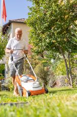 Elektro- vs. Akkurasenmäher: Der Vergleich Ein Mensch mit weißem T-Shirt mäht mit einem weiß-orangenen Akkurasenmäher seinen Garten. Die Sonne scheint und der Rasen und die Pflanzen sind grün.