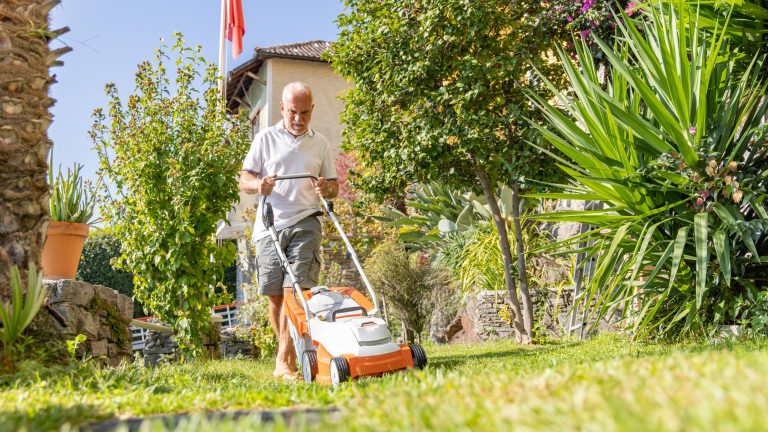 Ein Mensch mit wei&szlig;em T-Shirt m&auml;ht mit einem wei&szlig;-orangenen Akkurasenm&auml;her seinen Garten. Die Sonne scheint und der Rasen und die Pflanzen sind gr&uuml;n.
