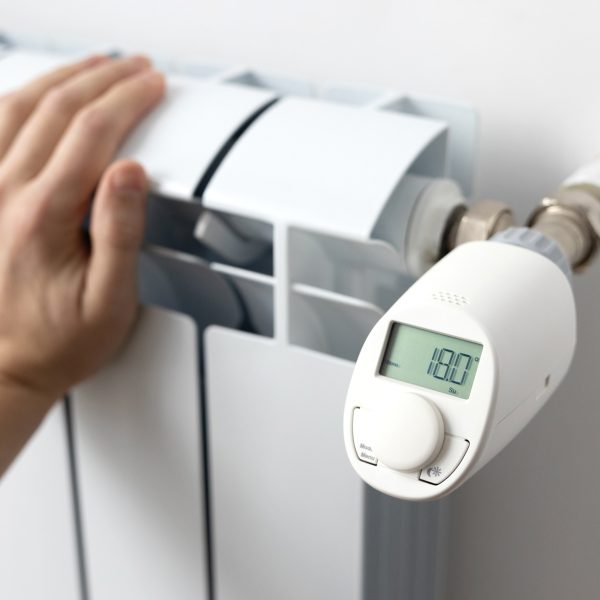 Eine Person fasst an eine Heizung, an der ein elektronisches Thermostat sitzt.