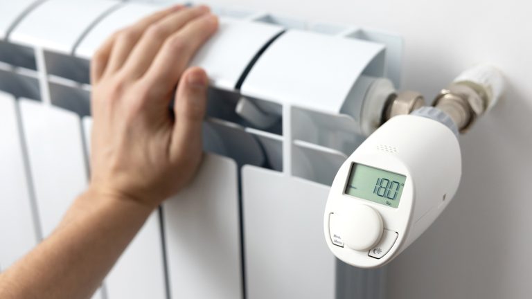 Eine Person fasst an eine Heizung, an der ein elektronisches Thermostat sitzt.