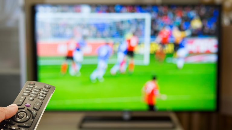 Eine Person hält eine Fernbedienung in der Hand und richtet sie auf einen Fernseher, auf dem ein Fußballspiel zu sehen ist.
