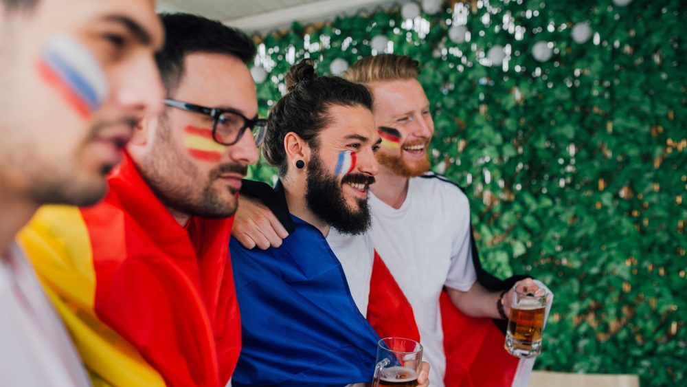 Vier Personen mit L&auml;nderfahnen auf die Wange geschminkt schauen in Richtung eines nicht sichtbaren Fernsehers.