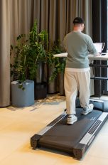 Eine Person steht in einem B&uuml;ro an einem Walking Pad, das unter einem Schreibtisch steht.