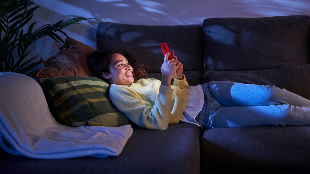Eine Person liegt im dunklen Wohnzimmer auf der Couch und scrollt an ihrem Handy.