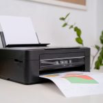 Ein Drucker steht neben einem Laptop und einer Pflanze auf einem Tisch und druckt ein Blatt mit bunten Mustern.