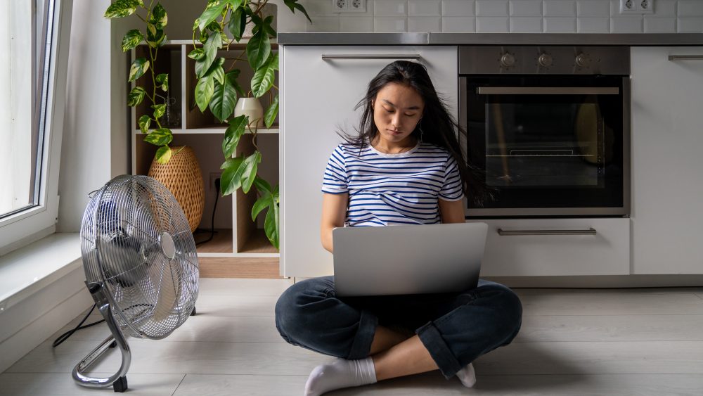 Eine Person sitzt auf dem Küchenboden angelegt an einen Schrank. Neben ihr steht ein klassischer Ventilator.