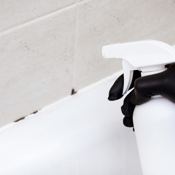 Eine Person h&auml;lt eine Spr&uuml;hflasche in der Hand, die in Richtung von Schimmel an Silikonfugen gerichtet ist.