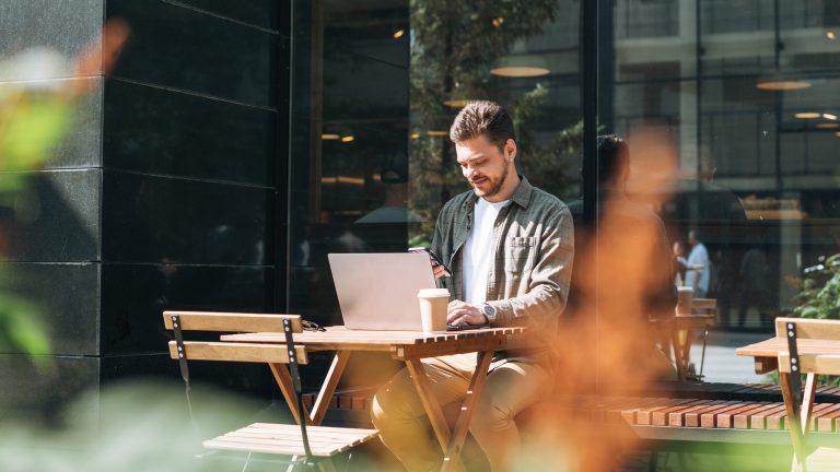 Eine Person sitzt vor einem Caf&eacute; an einem Tisch und arbeitet mit einem Laptop und einem Smartphone.