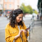 Eine Person ist im Freien unterwegs, tr&auml;gt Kopfh&ouml;rer und bedient dabei ihr Smartphone.