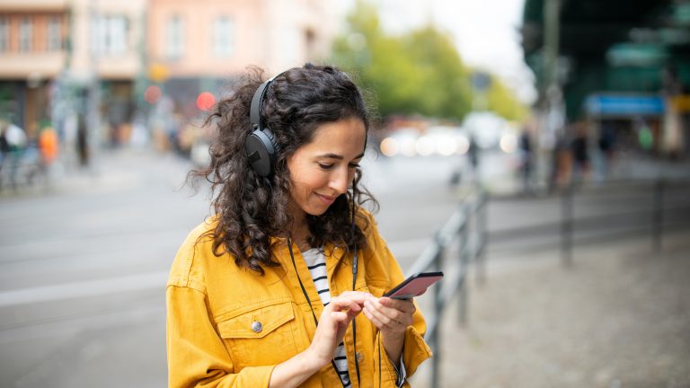 Eine Person ist im Freien unterwegs, trägt Kopfhörer und bedient dabei ihr Smartphone.