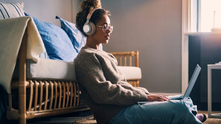 Eine Person sitzt vor einem Sofa auf dem Boden, trägt Kopfhörer und hat einen Laptop auf dem Schoß.