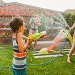 Eine Person h&auml;lt einen Gartenschlauch, aus dem Wasser in Richtung eines Kindes spritzt, das eine Wasserpistole in der Hand h&auml;lt.
