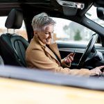 Eine Person sitzt in einem Auto und tippt auf dem Infotainment-System des Autos herum. In der anderen Hand h&auml;lt sie ein Smartphone.