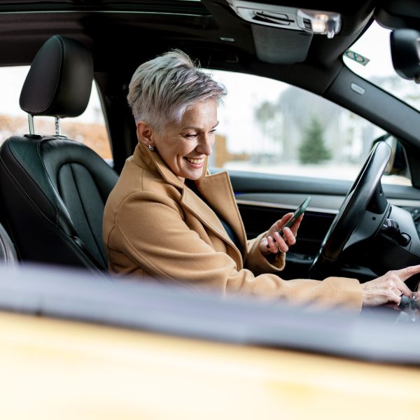 Eine Person sitzt in einem Auto und tippt auf dem Infotainment-System des Autos herum. In der anderen Hand h&auml;lt sie ein Smartphone.