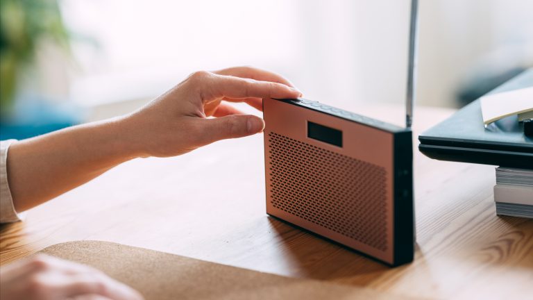 Eine Person drückt einen Knopf an der Oberseite eines schmalen, kleinen Radios auf einem Schreibtisch. 