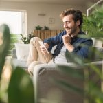 Eine Person sitzt in einem Wohnzimmer mit vielen Pflanzen auf der Couch und scrollt auf ihrem Smartphone.