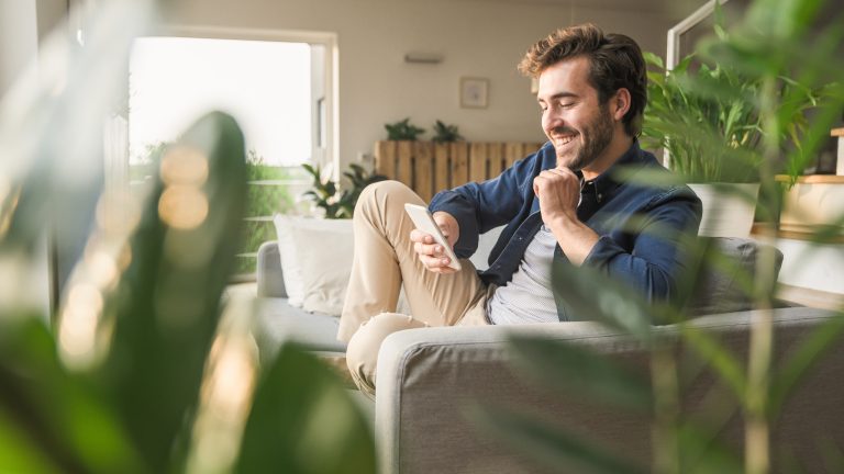 Eine Person sitzt in einem Wohnzimmer mit vielen Pflanzen auf der Couch und scrollt auf ihrem Smartphone.