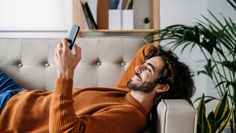 Eine Person liegt in einem Wohnzimmer auf der Couch und scrollt auf ihrem Smartphone.