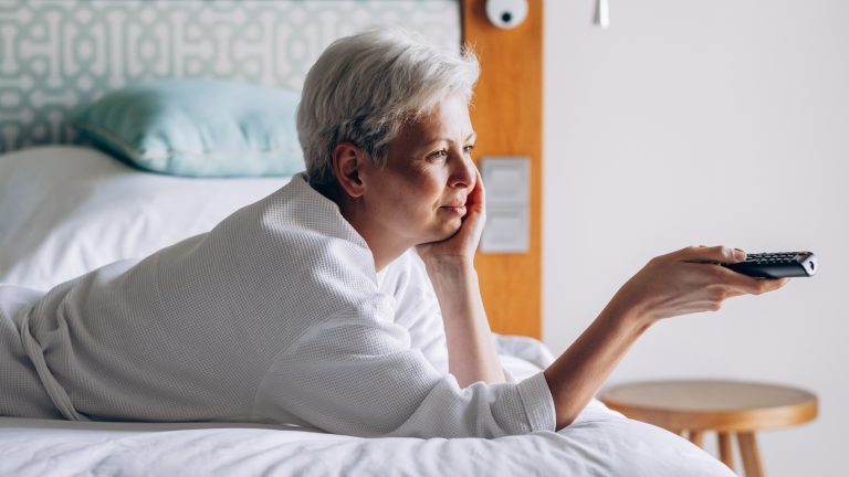 Eine Person liegt b&auml;uchlings auf einem Bett und richtet eine Fernbedienung in die Ferne.