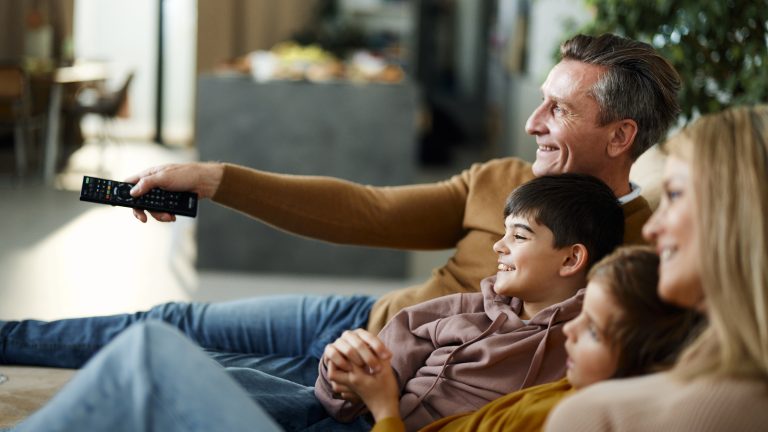 Zwei Personen und zwei Kinder sitzen nebeneinander auf der Couch im Wohnbereich. Eine Person richtet eine Fernbedienung auf etwas in der Ferne.